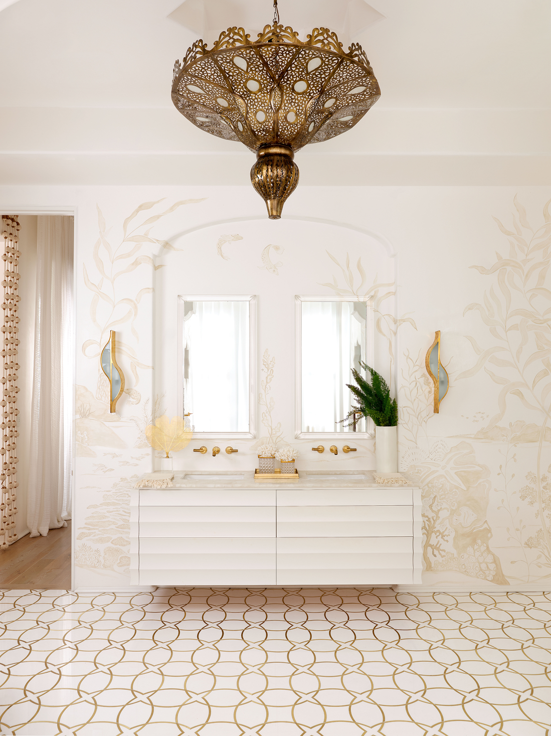 A white bathroom with gold accents, featuring a double vanity, two mirrors, a decorative chandelier, and beige botanical wall art.