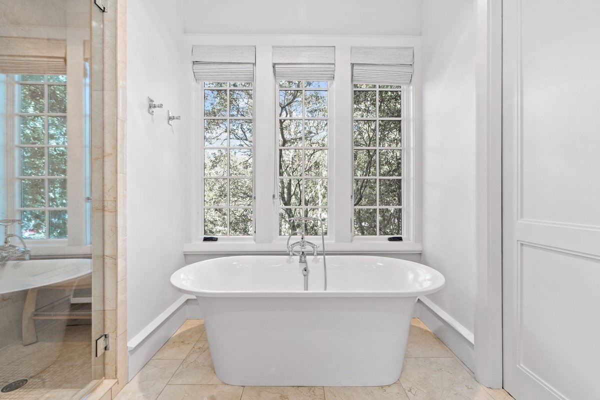 Bathroom with a white bathtub in front of three large windows with white blinds, beige tiled floor, and a partial view of the shower on the left.