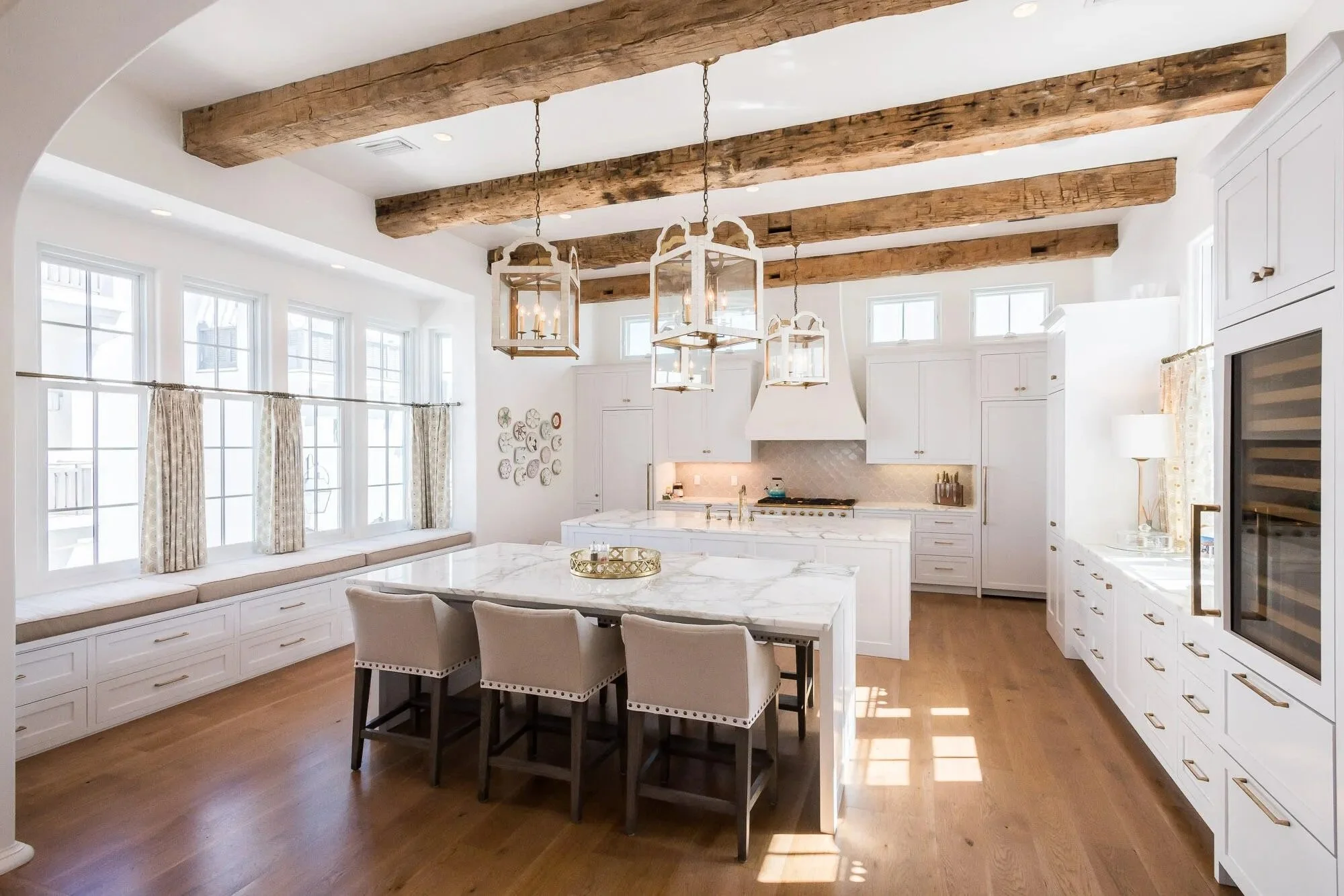 Bright kitchen with white cabinetry, wooden ceiling beams, large window seat, marble island, and hanging lantern light fixtures.