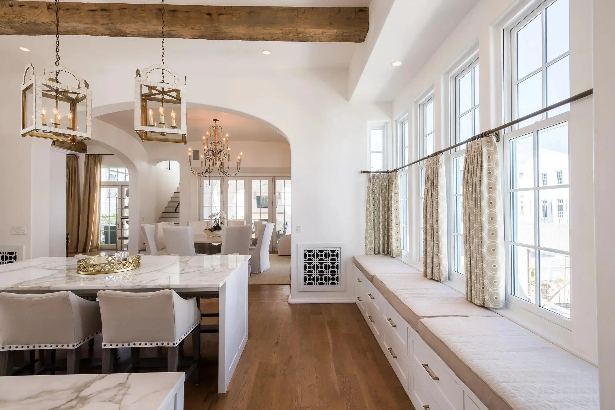 Bright living space with large windows, a built-in window seat with drawers, and a dining area with a chandelier in the background.