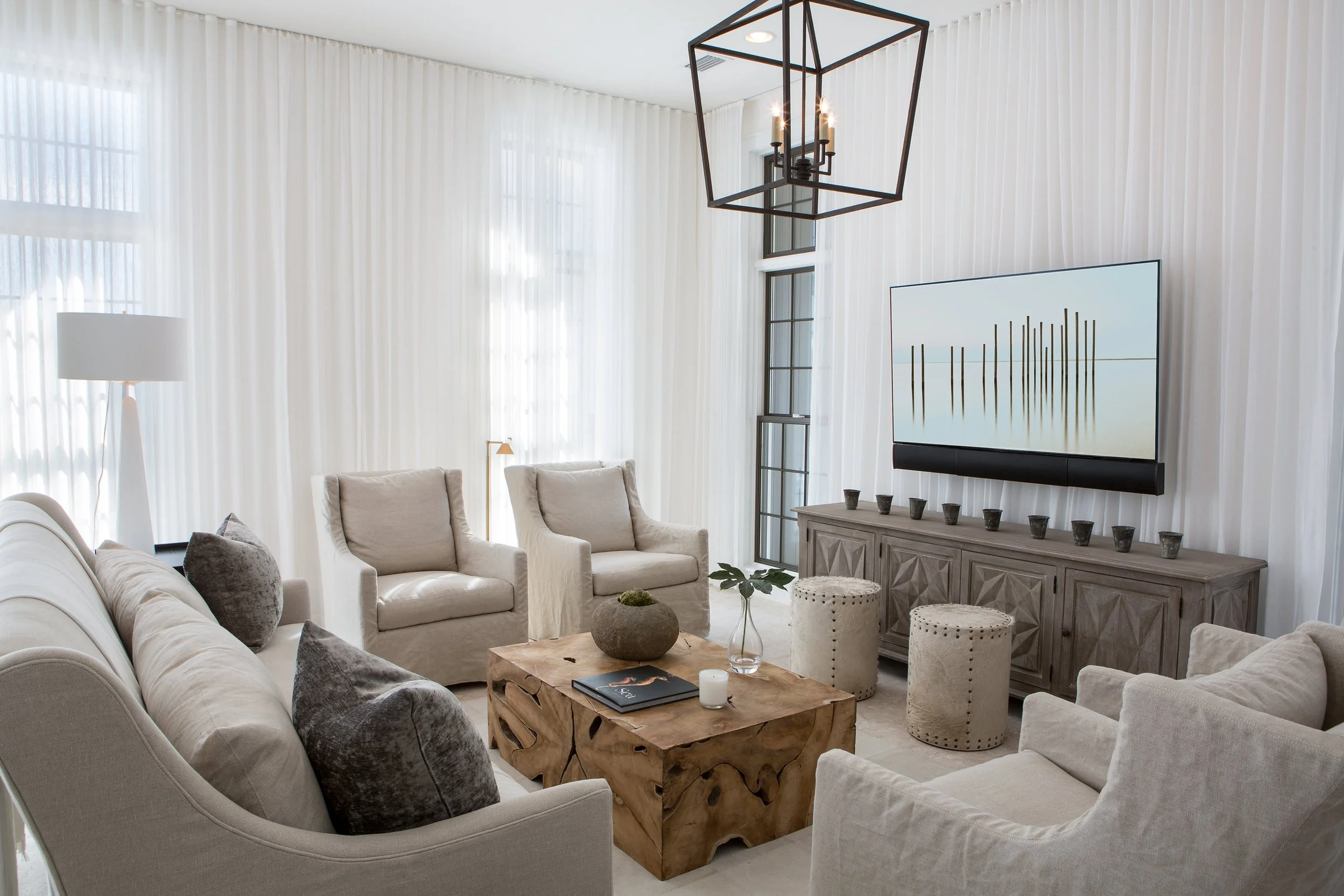 Living room with white walls and sheer curtains, beige sofas and armchairs, wooden coffee table, wall-mounted TV, decorative stools, and a geometric chandelier.
