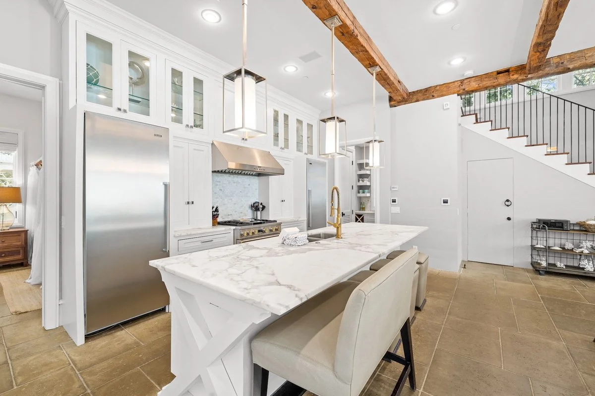 Modern kitchen with white cabinets, marble island, stainless steel appliances, wooden beams on the ceiling, and beige tile flooring.