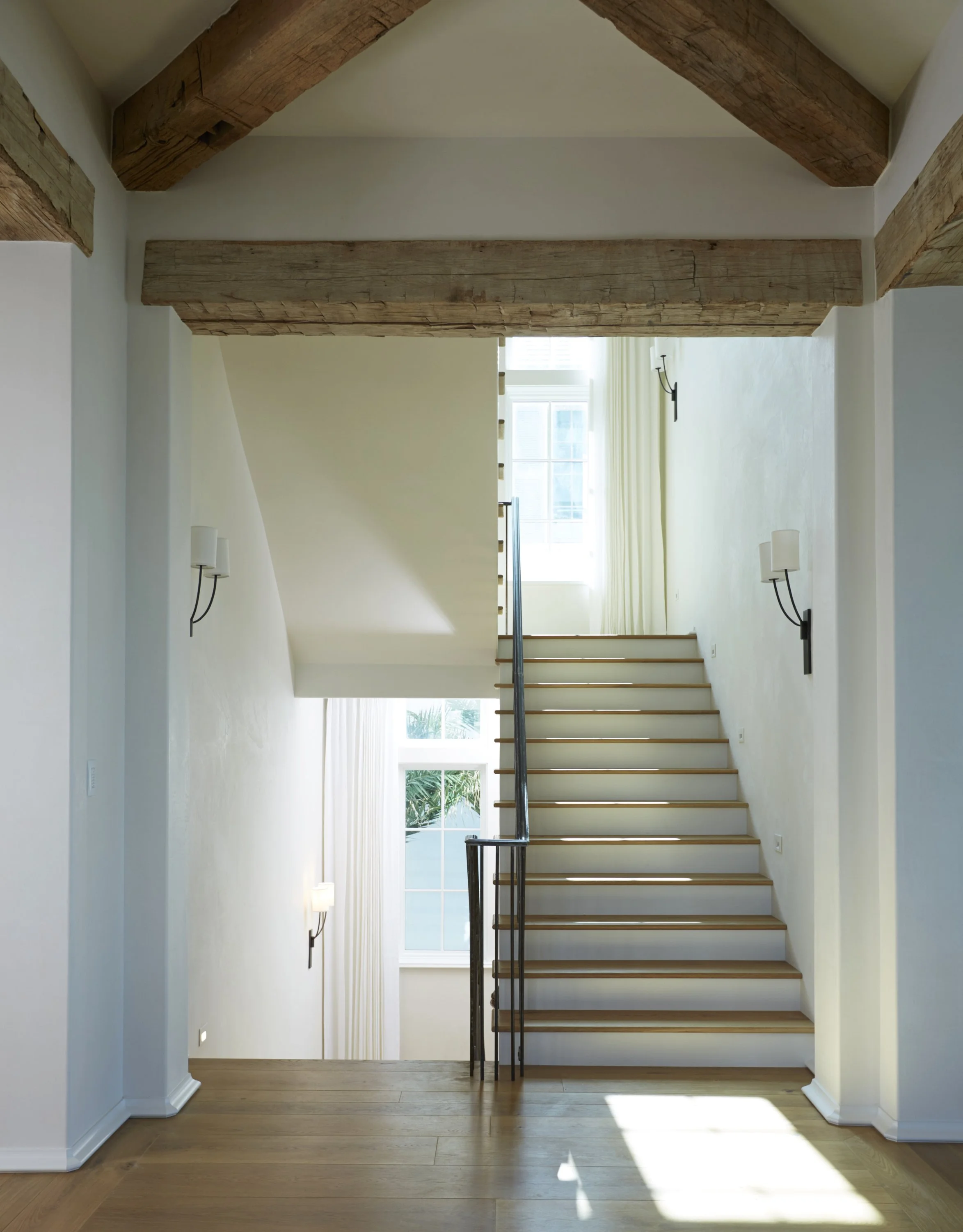 Interior view of a modern staircase with wooden steps, black metal railing, and large windows letting in natural light. Exposed wooden beams and wall-mounted lights are visible.