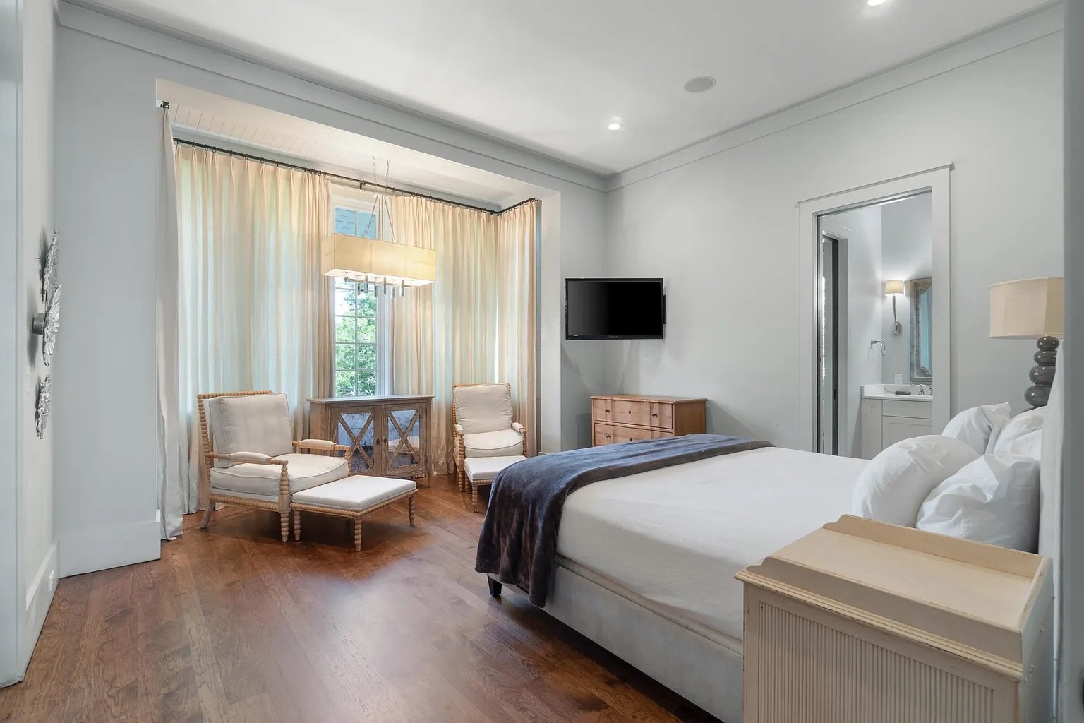 A bedroom with a large bed, white bedding, and pillows, a nightstand, a wall-mounted TV, two chairs near a window, and a bedside lamp, with a bathroom visible in the background.