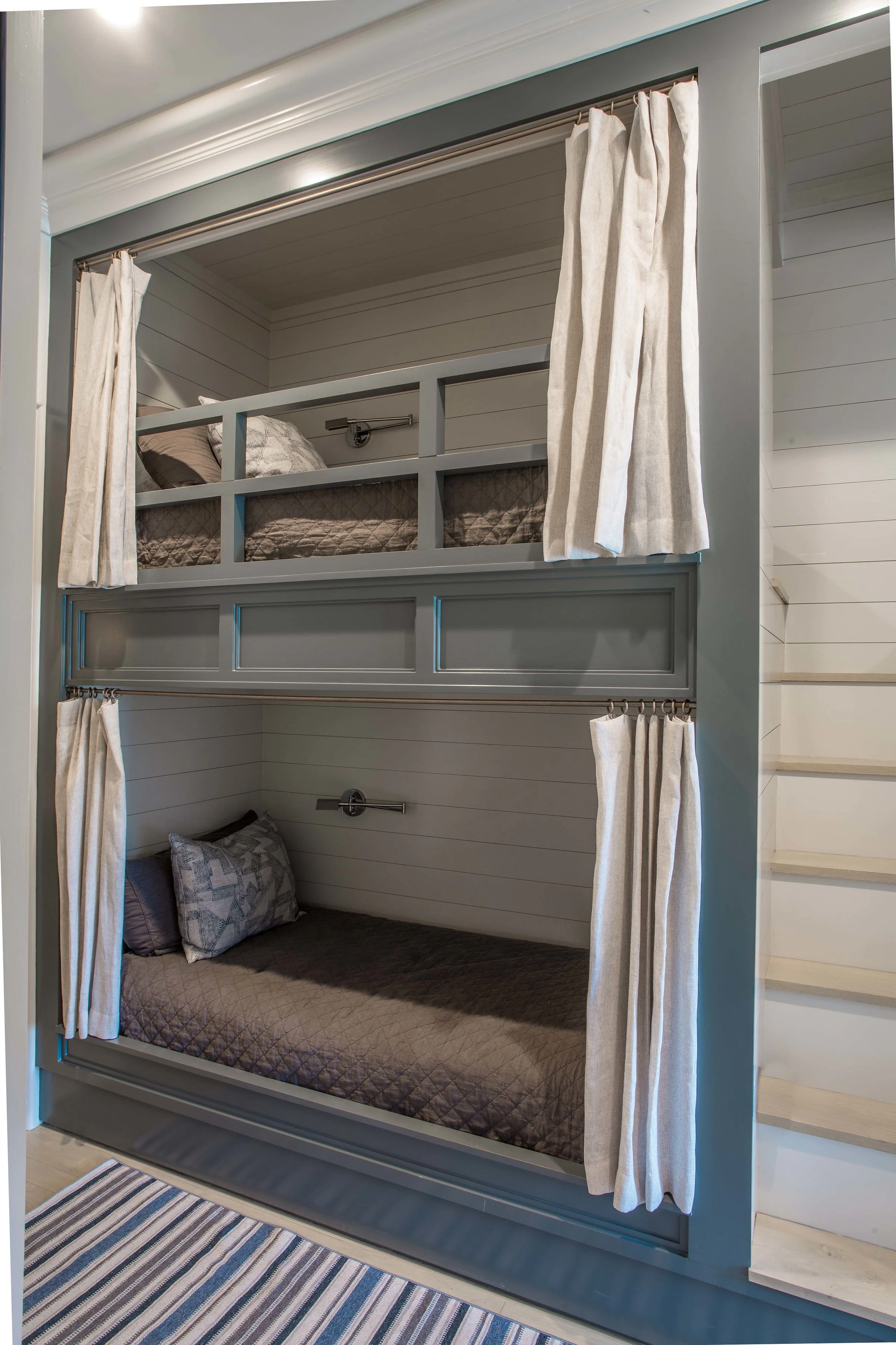 Bunk beds with curtains for privacy in a bedroom, with the top bunk featuring a bed with gray bedding and the bottom bunk having a bed with dark gray bedding and pillows. There are wall-mounted reading lights above each bed.