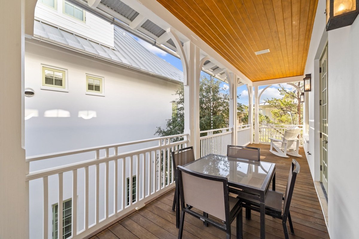Covered porch with a glass-top table and four chairs, white rocking chair, wooden ceiling, white railing, wall-mounted lantern, and view of trees and neighboring house.
