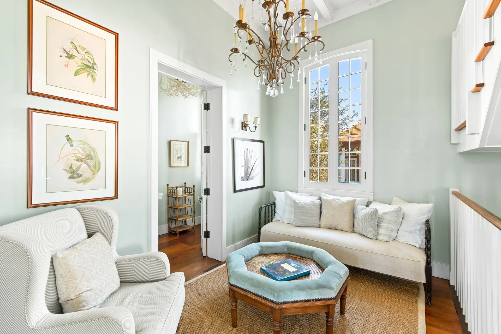 A cozy living room with pale green walls, an elegant chandelier, a window seat with beige and white pillows, artwork on the walls, and a hardwood floor with a woven rug.