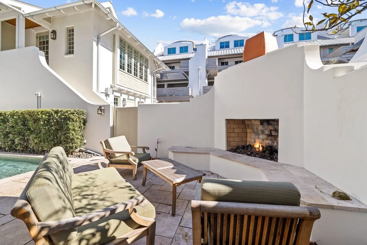 A cozy outdoor seating area with a fireplace in a residential backyard, featuring a sofa, chairs, a small table, and a hedge, with multi-story buildings in the background.