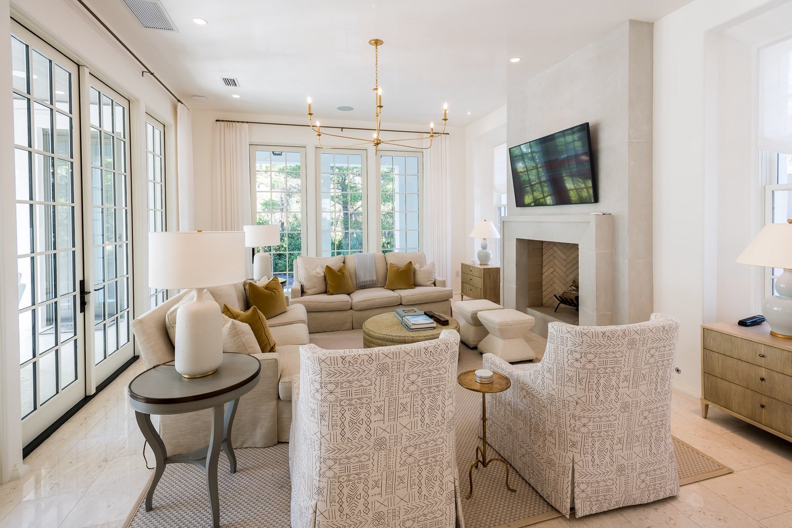 Bright living room with cream-colored sofas, patterned armchairs, a fireplace, large windows providing natural light, and a mounted flat-screen TV.