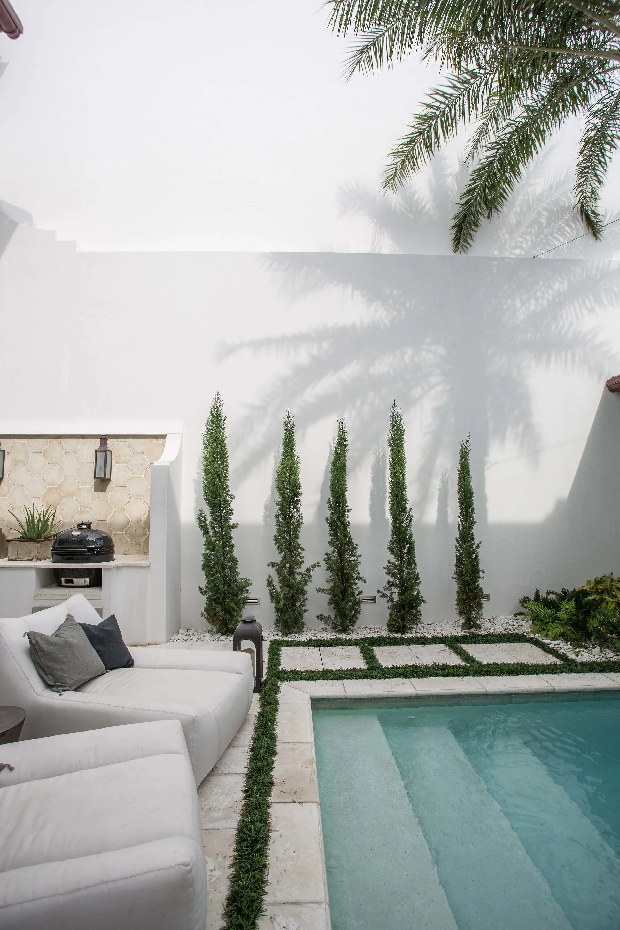 A modern backyard with a swimming pool, white outdoor furniture, and a white wall with tall, narrow trees and palm tree shadows.