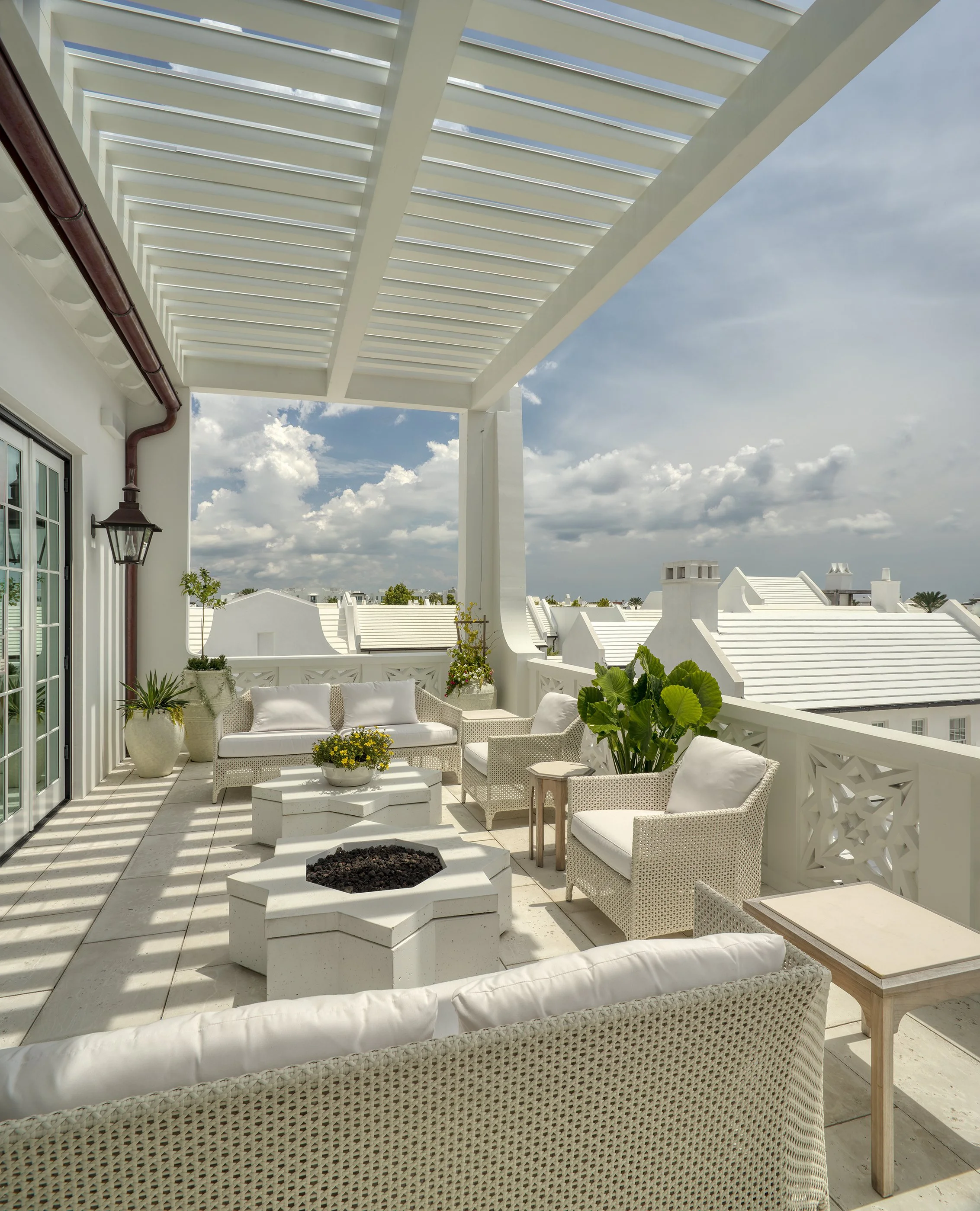 Sunlit outdoor balcony with white wicker furniture, potted plants, and a fire pit, overlooking a neighborhood with white roofs under a partly cloudy sky.