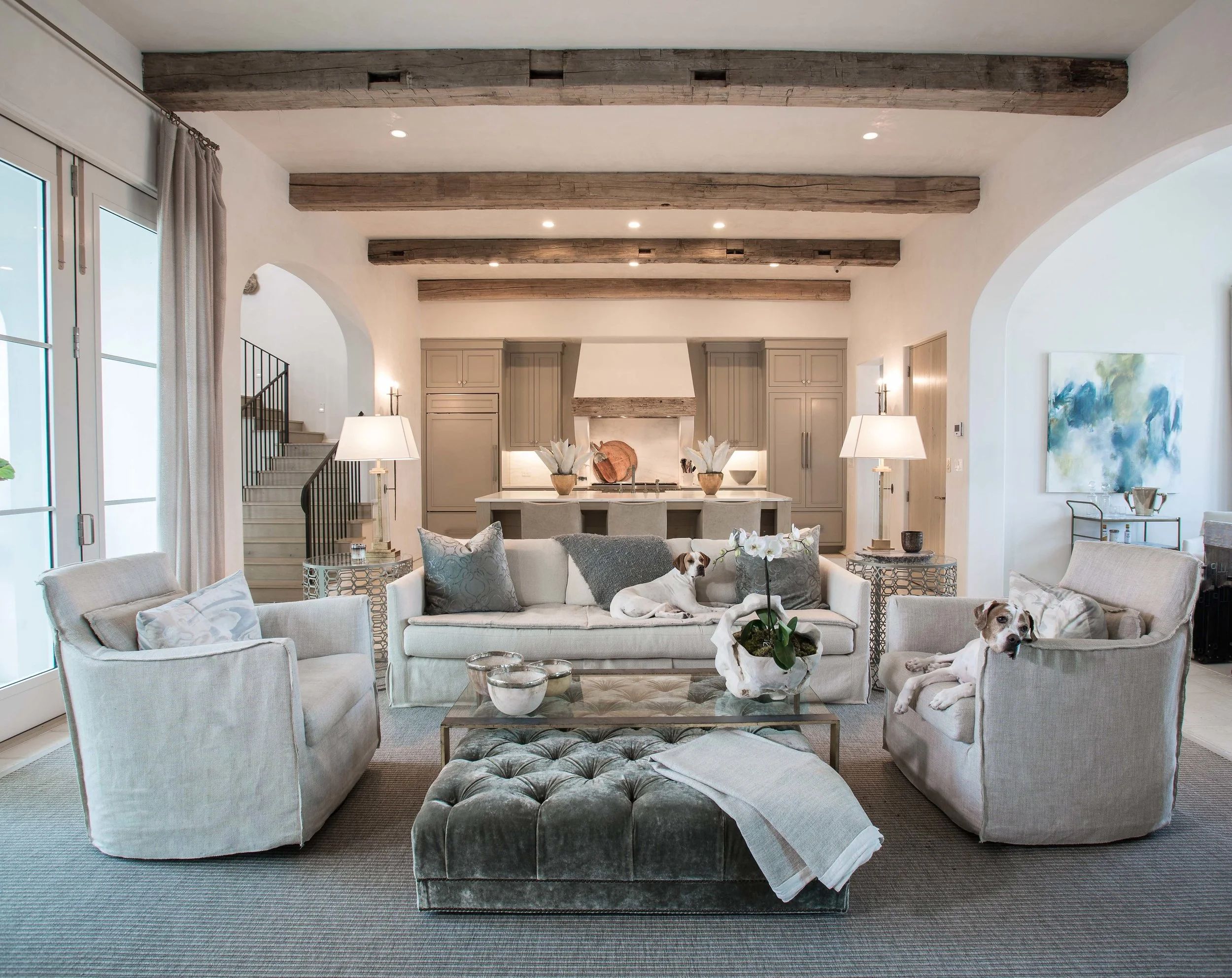 Elegant living room with white and beige furniture, decorative pillows, coffee table, and dogs lounging on sofas. Open kitchen in the background with cabinetry, pendant lighting, and a staircase to the left. Wooden ceiling beams add rustic charm.