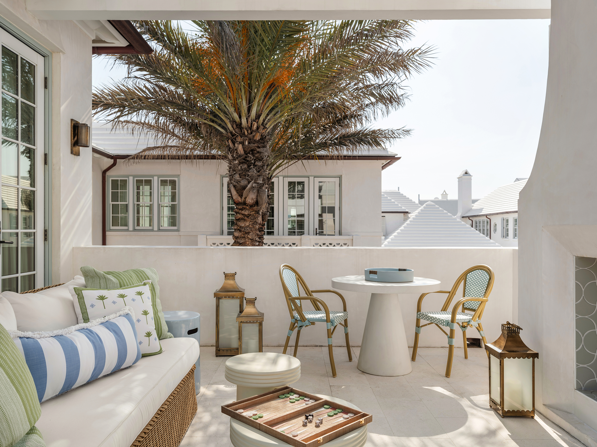 Outdoor patio with a white sofa with green and blue pillows, a round white table with two chairs, a large palm tree, and decorative lanterns, under a covered area with a view of neighboring rooftops.