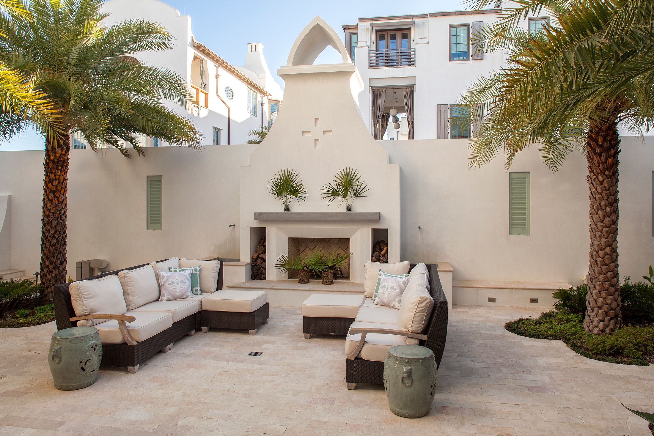 An outdoor patio with white cushioned seating, two ceramic stools, palm trees, and a white stucco building with a decorative fireplace.