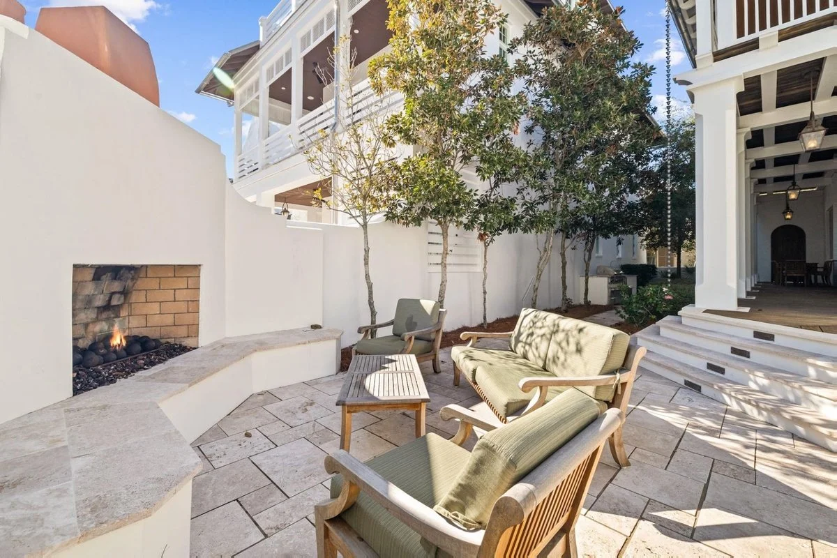 Outdoor patio area with a white curved fire pit, three wooden benches with green cushions, trees, and stairs leading up to a house with a porch.