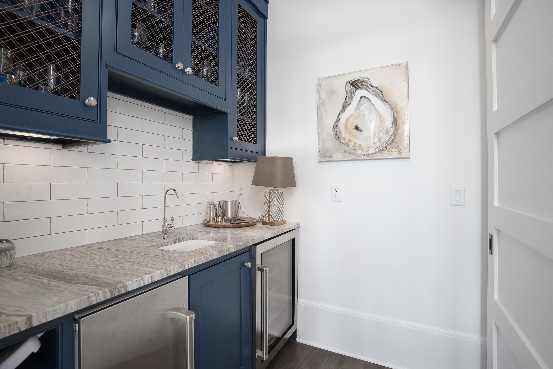 A compact wet bar area with blue cabinetry and a granite countertop, small sink, and a wine cooler. Decor includes a table lamp and abstract artwork on a white wall.