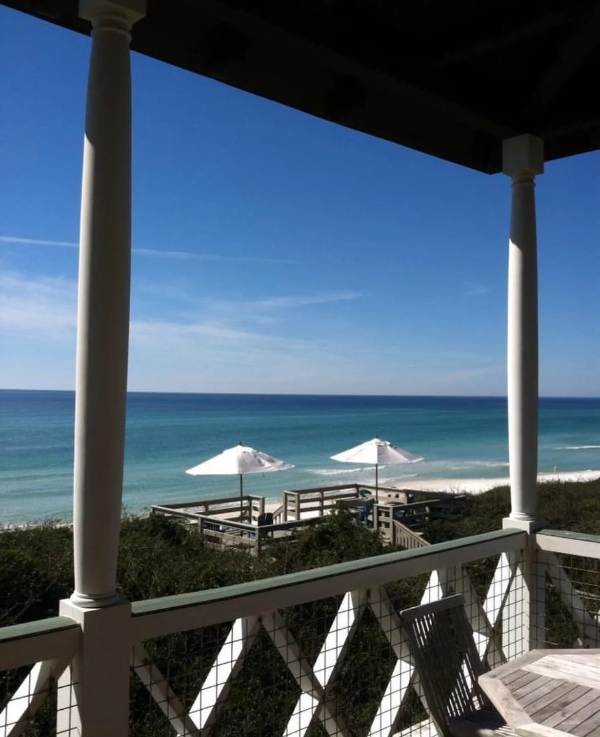 View from a balcony overlooking a beach with white umbrellas, wooden deck, and blue ocean under a clear sky.