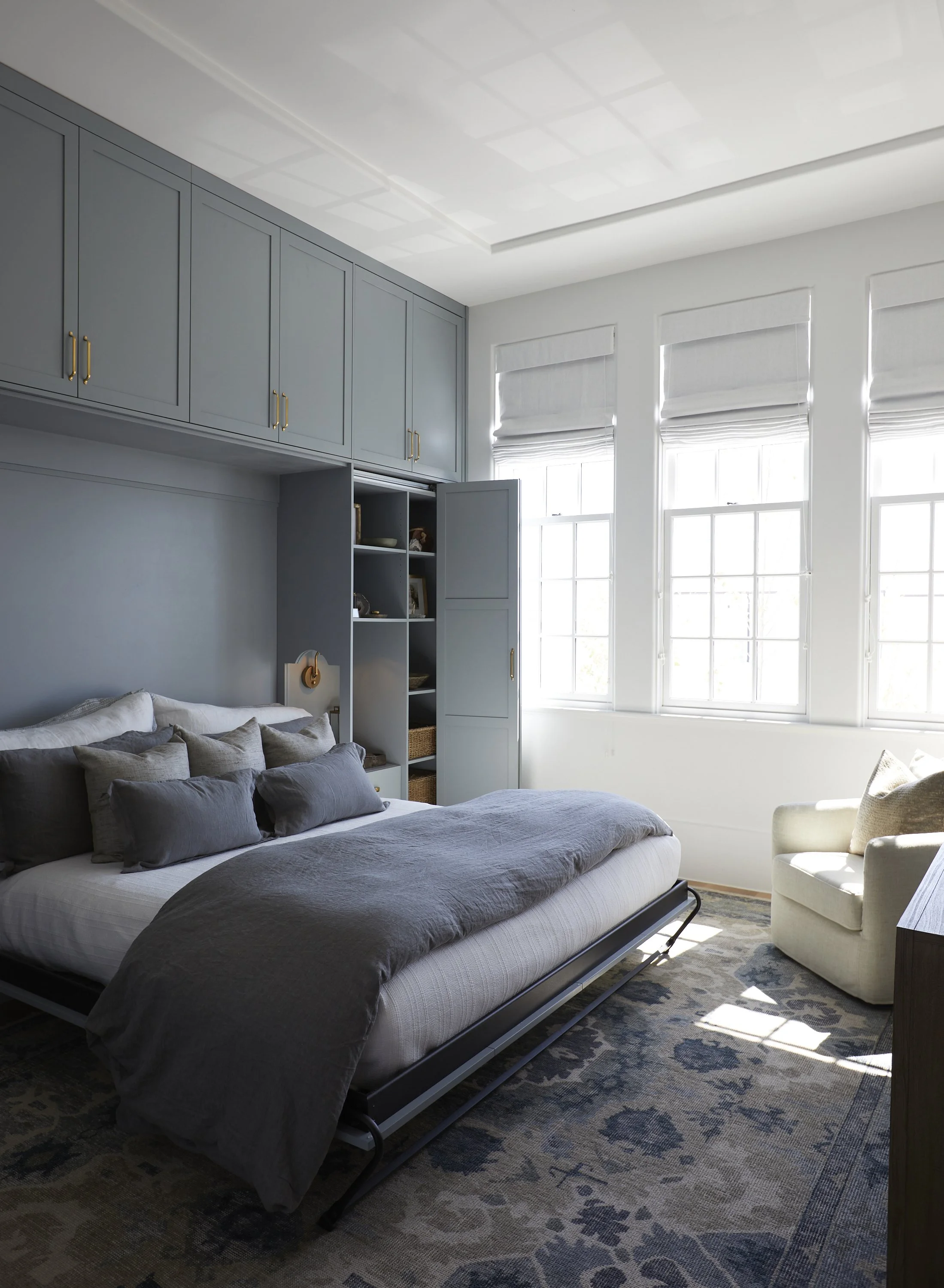 A bedroom with large windows, a bed with gray bedding, a white armchair, built-in blue cabinets, and a patterned area rug.