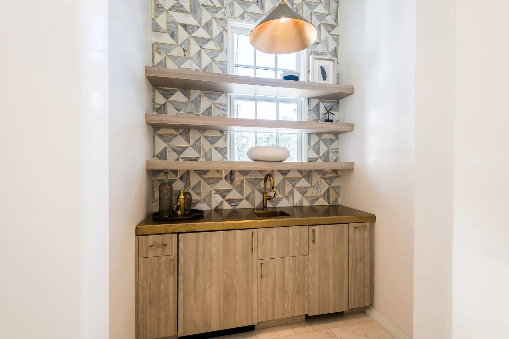 A small kitchen or bar area with a wooden cabinet, a black countertop, a gold faucet, and three open wooden shelves on a geometric patterned tile backsplash. There are decorative vases and a window with blinds behind the shelves, and a rose gold pend