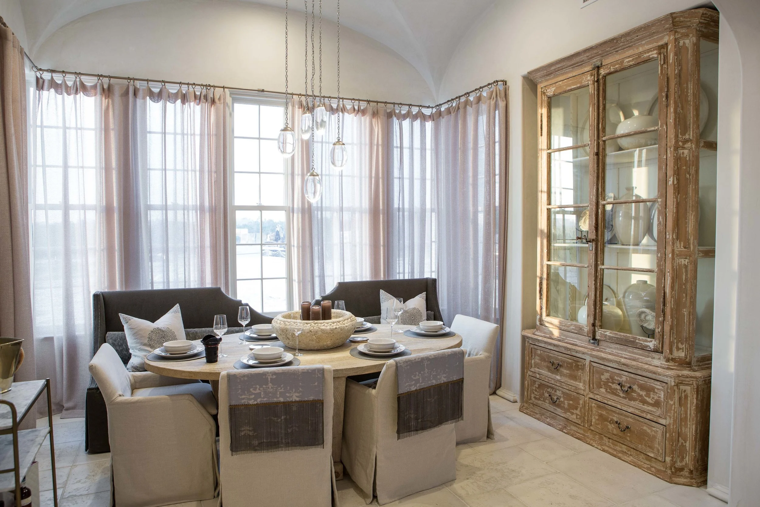 Dining room with a round table set for dining, surrounded by cushioned chairs, with large windows, sheer curtains, a vintage wooden display cabinet, and modern pendant lighting.