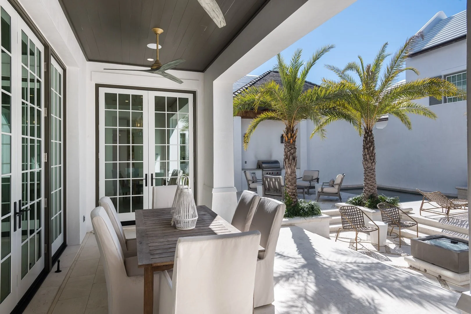 Outdoor patio with a dining table and chairs, palm trees, and lounge seating, with a barbecue grill in the background under a clear blue sky.