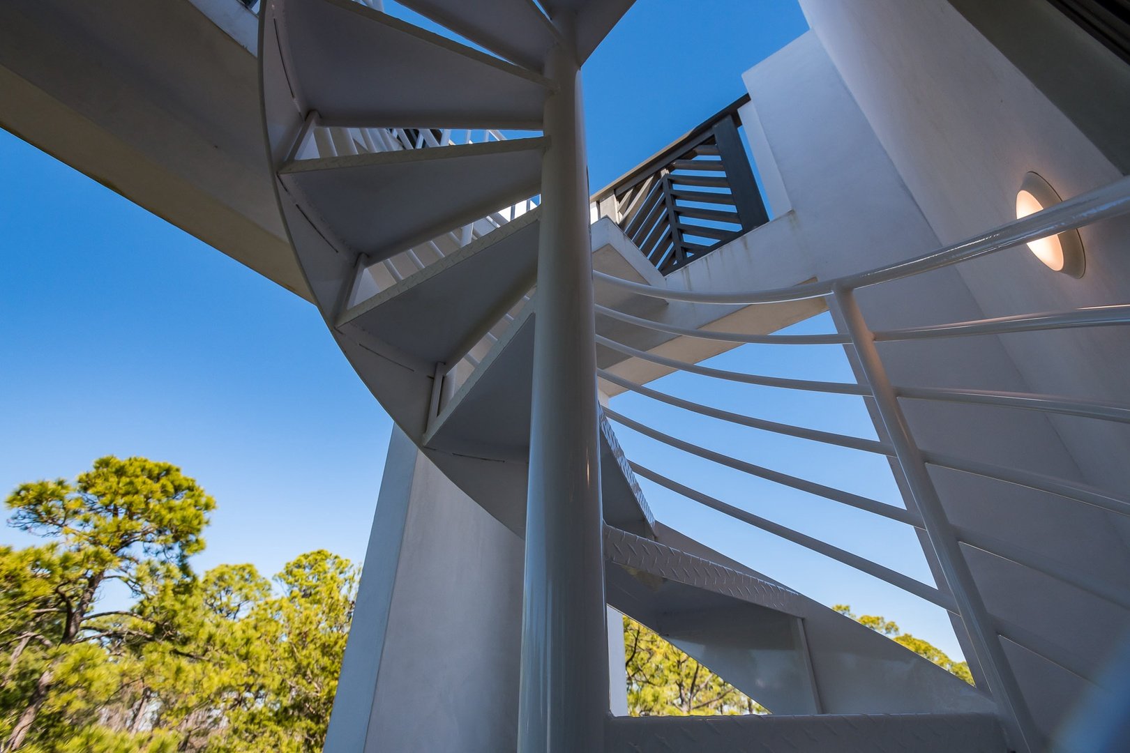 Interior view of a white spiral staircase outdoors with trees and a blue sky in the background.