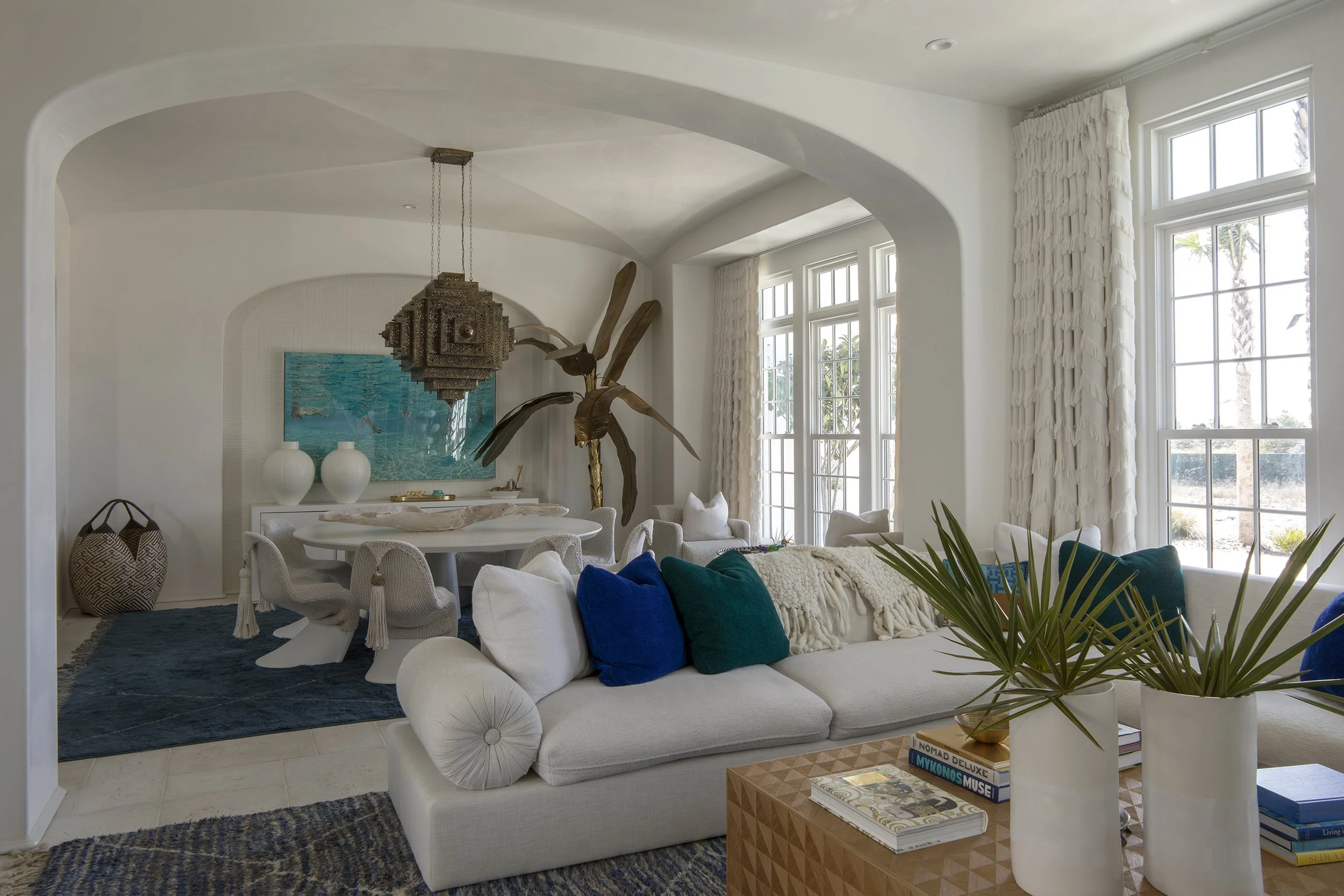 Bright living room with large windows, white sofa with blue and green pillows, decorative plant on a wooden table, and a dining area with white table and chairs in the background.