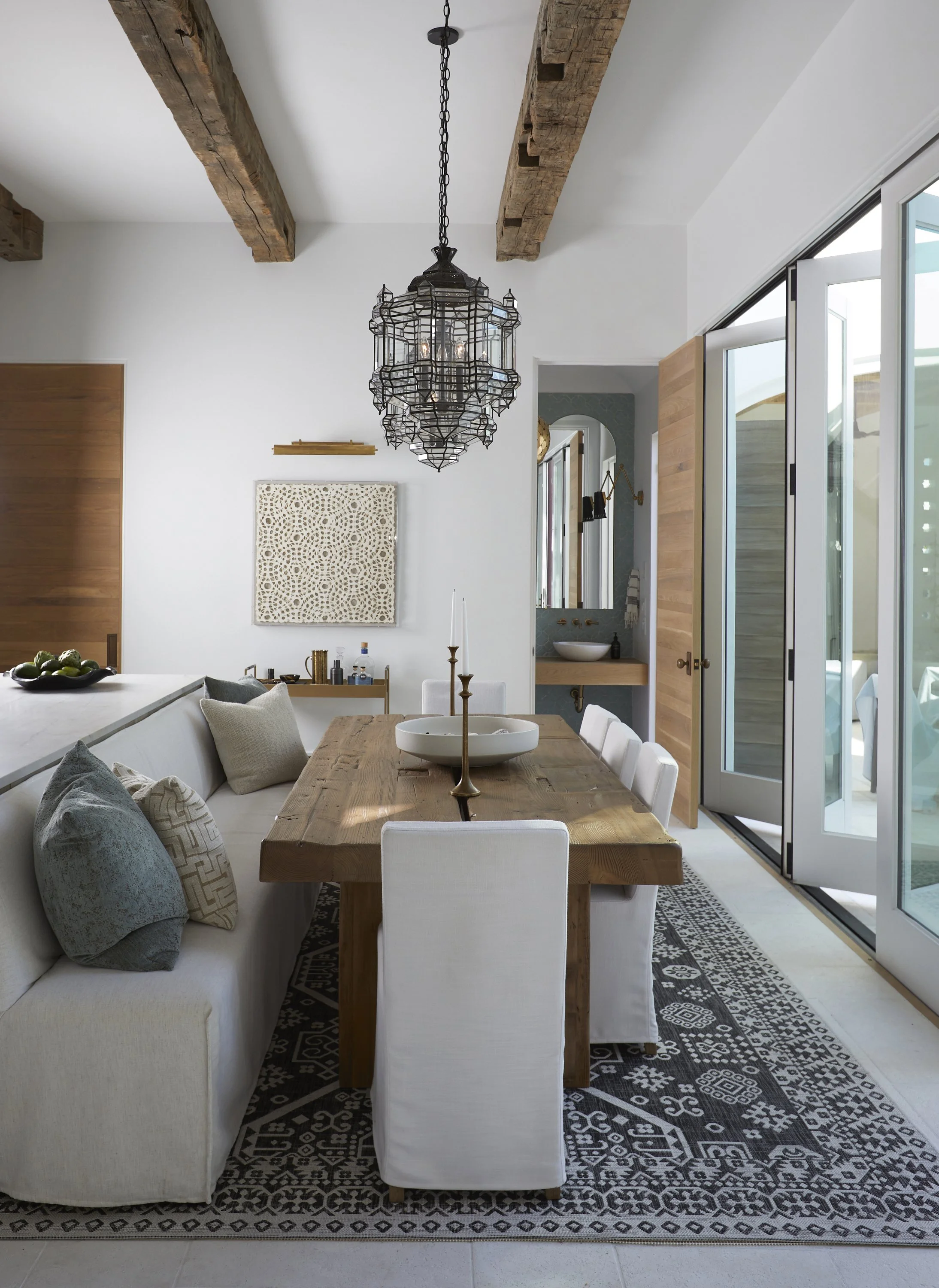 Modern dining room with wooden table, white chairs, and a built-in white sofa with pillows, decorated with candles, artwork, and a chandelier, with sliding glass doors leading outside.