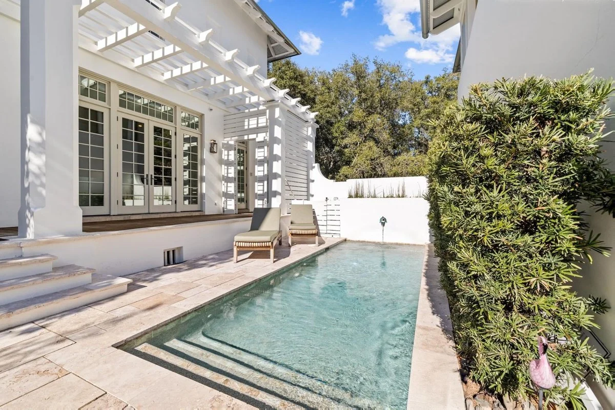 A backyard with a small pool, two lounge chairs, a white wooden pergola, and lush green bushes against a blue sky.
