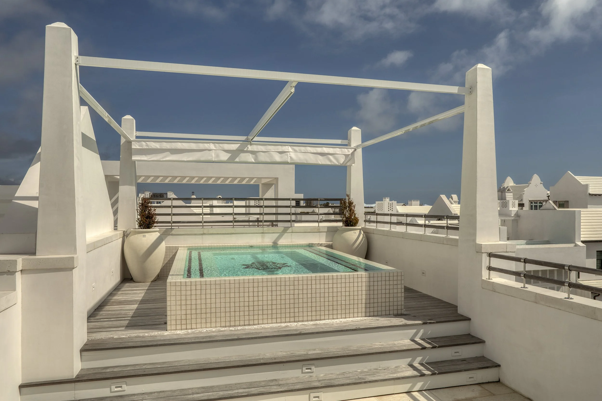 White rooftop terrace with small swimming pool, potted plants, and a white pergola against a partly cloudy sky.