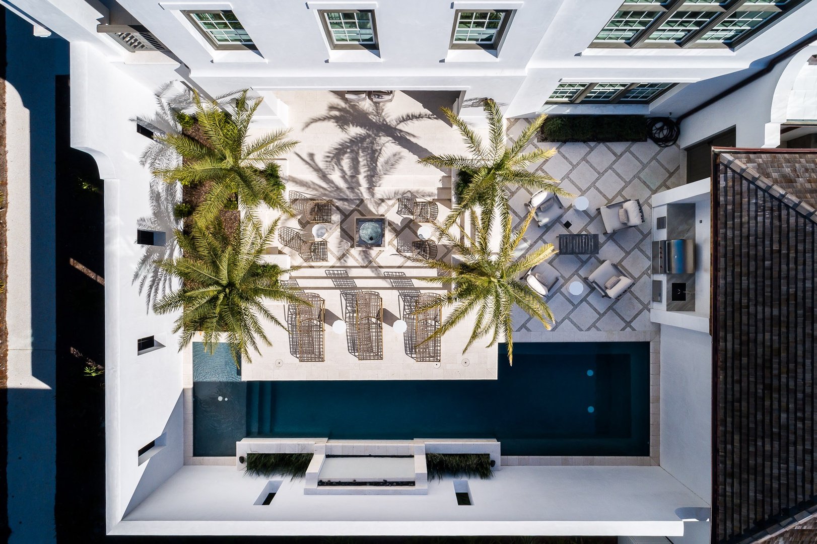 Aerial view of a modern outdoor patio with a swimming pool, lounge chairs, palm trees, and seating areas, surrounded by a white building with multiple windows.