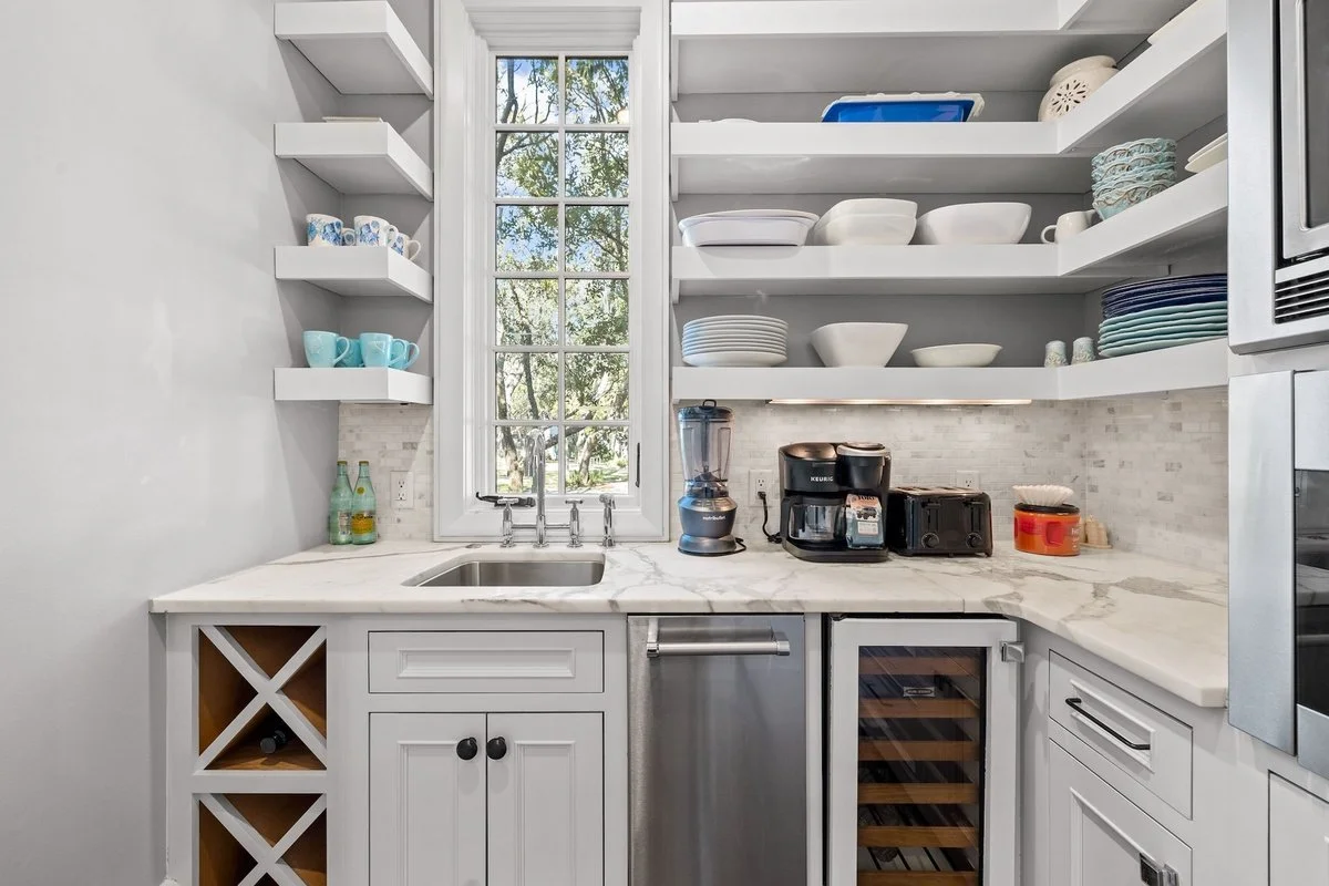 Kitchen with white shelves holding dishes and cups, a window behind the sink, stainless steel appliances, and a marble countertop.