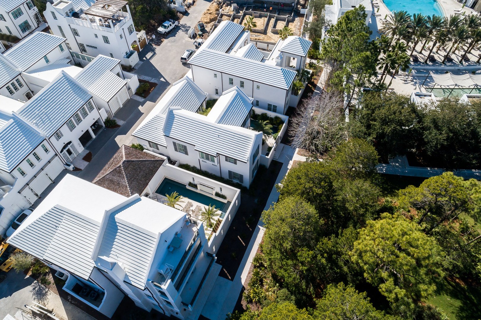 Aerial view of a modern residential neighborhood with white houses featuring metal roofs, a swimming pool, and large trees.
