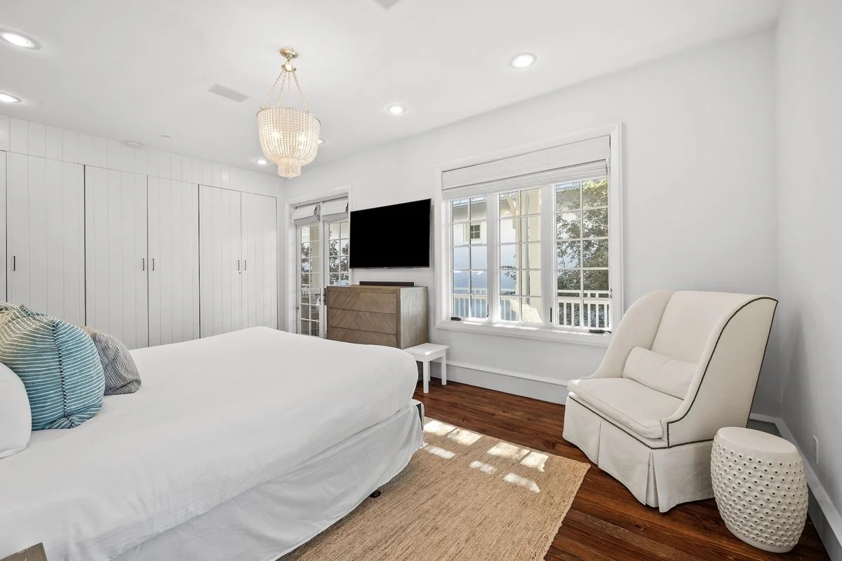 Bright bedroom with white walls, a large bed with a white bedspread and pillows, a white armchair, a round side table, a flat-screen TV on a chest of drawers, a large window with white blinds, and a closet with white doors.