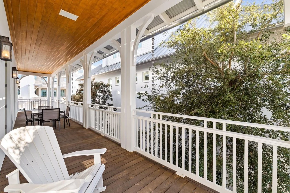 View of a spacious porch with wooden flooring, white railing, and white Adirondack chair. The porch has hanging lantern lights and outdoor tables and chairs, with a large tree on one side and neighboring houses in the background.