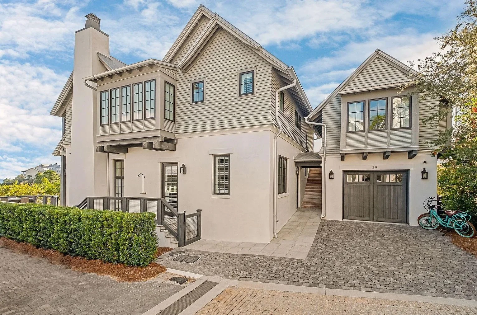 Front view of a modern multi-story house with beige siding and a gray garage door, surrounded by cobblestone and paved walkways, green bushes, two bicycles, and a partly cloudy sky.