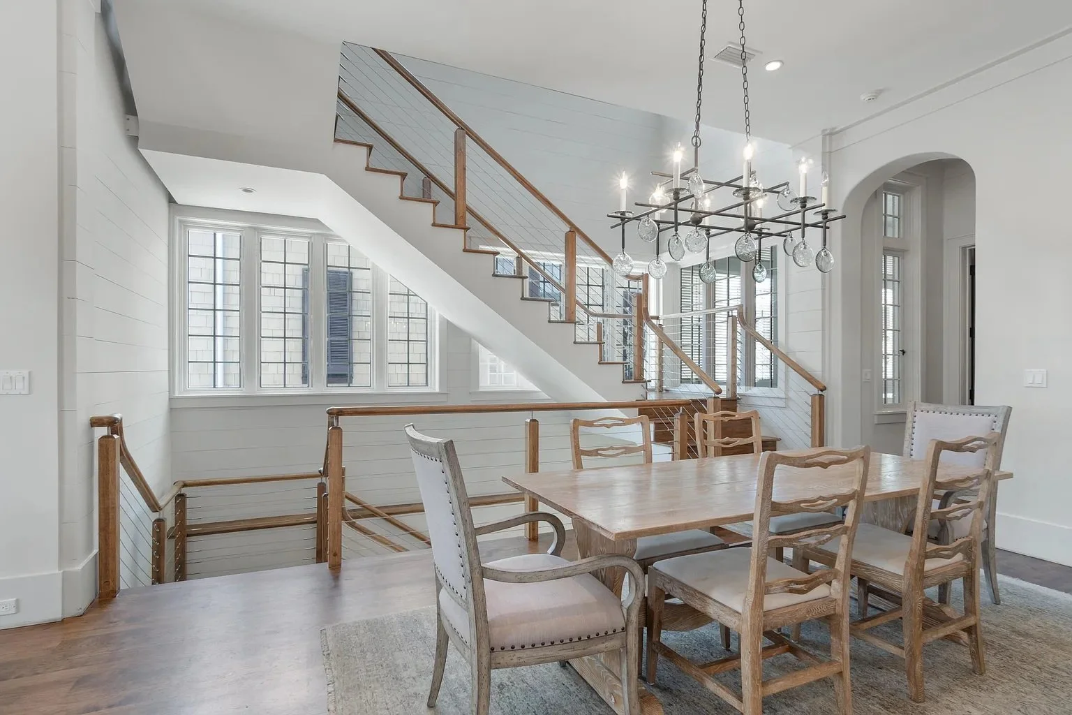 Bright dining room with wooden table and chairs, large windows, white walls, and a modern chandelier with candles and glass orbs.