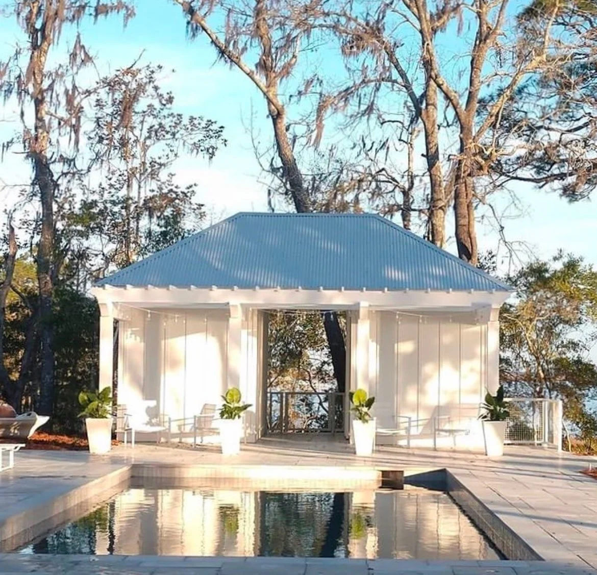 White gazebo with a blue metal roof, potted plants, and lounge chairs on a patio with a swimming pool, surrounded by leafless trees and blue sky.