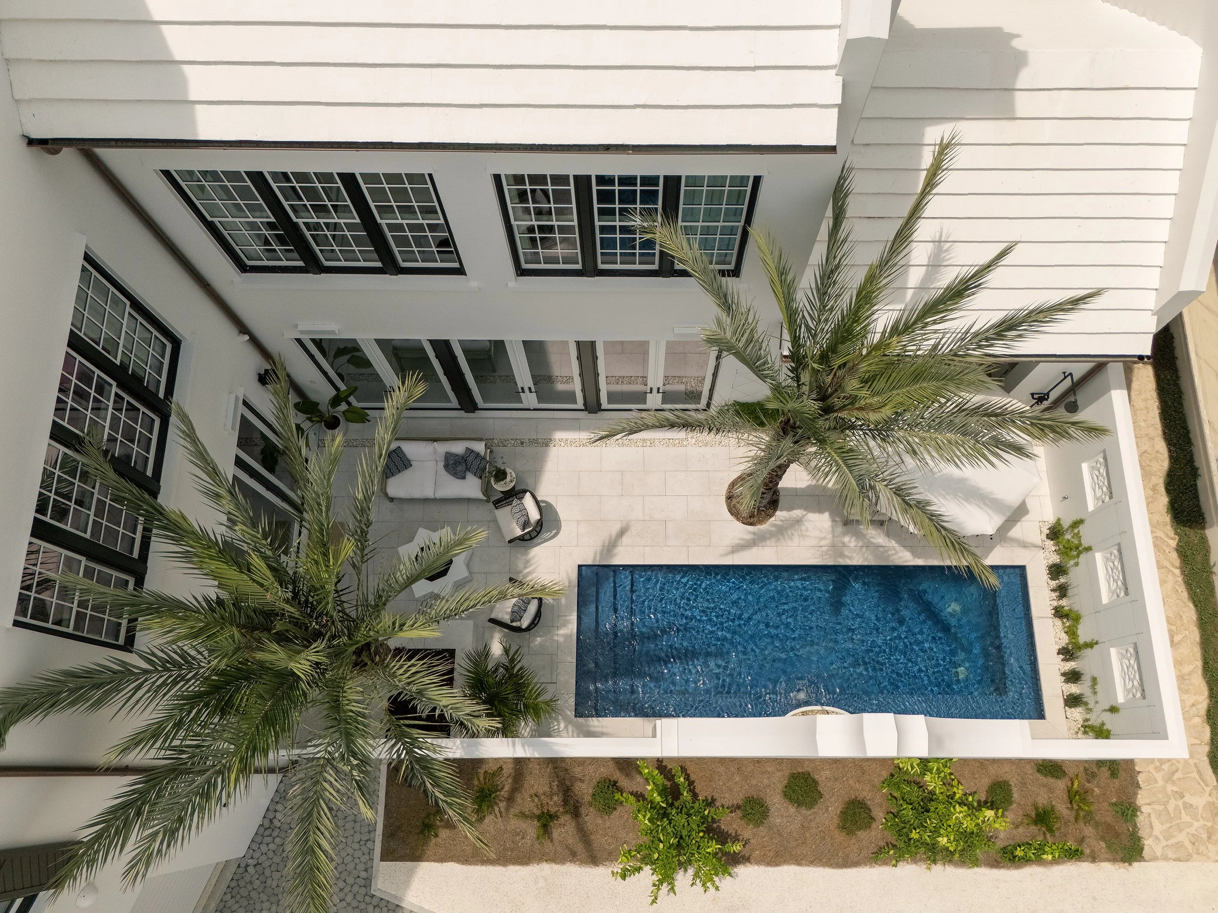 Aerial view of a modern backyard with a small swimming pool, palm trees, outdoor seating, and patio area surrounded by white walls and house windows.