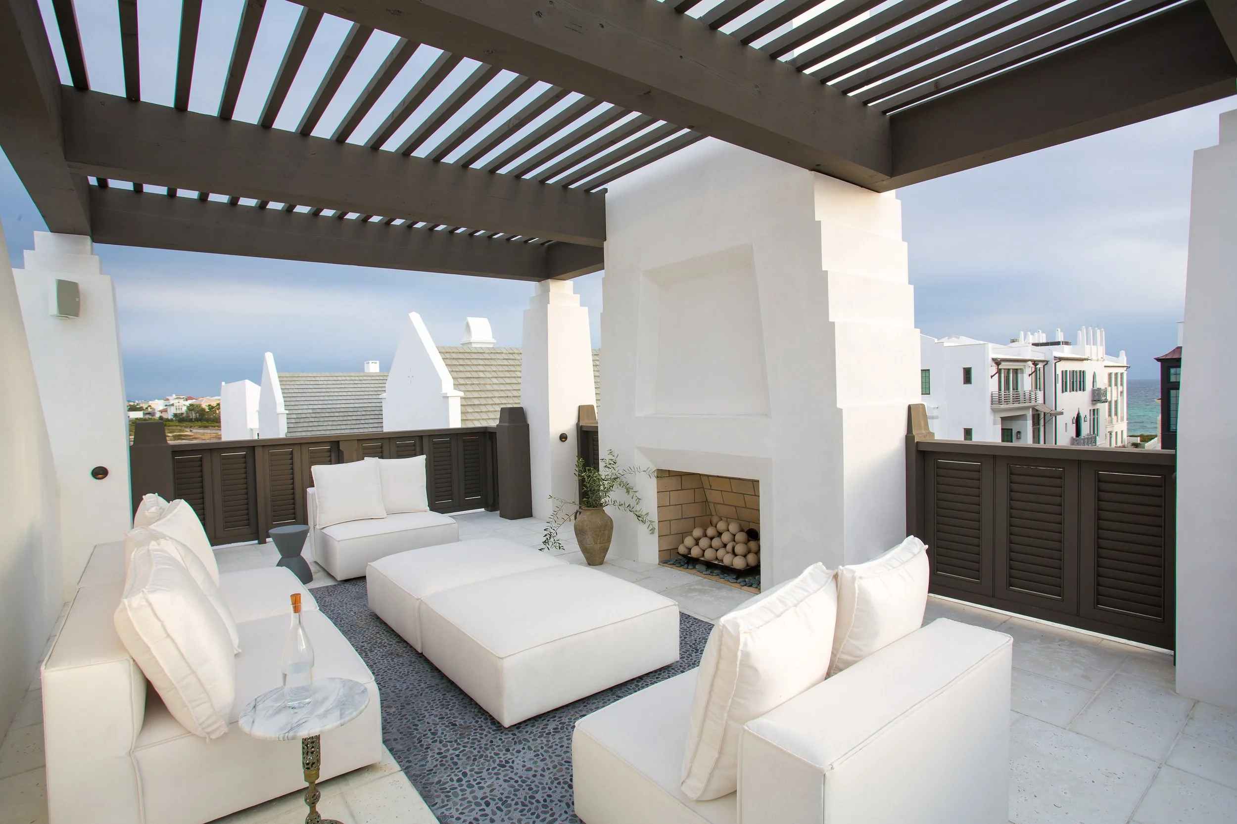 Outdoor rooftop balcony with white seating, a fireplace, and a view of neighboring buildings with rooftops and the ocean in the distance.