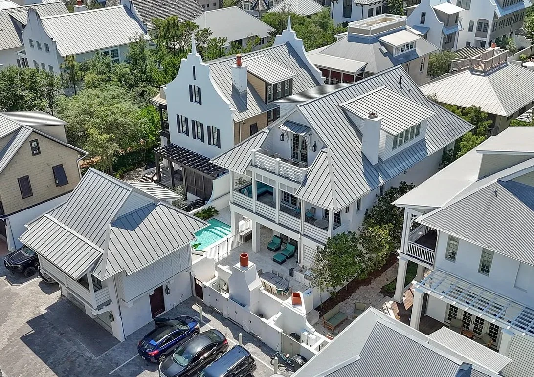 Aerial view of a neighborhood with white and beige houses featuring metal roofs, a backyard with a small pool, outdoor seating area, and surrounding greenery.
