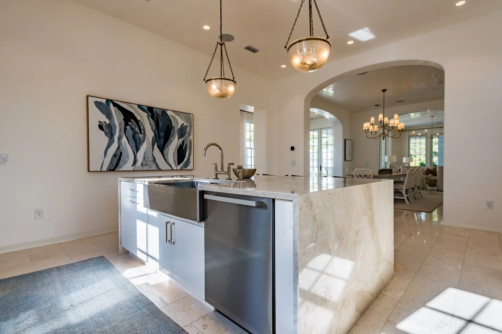 Modern kitchen with a marble island, stainless steel sink, pendant lights, artwork on the wall, and a view into a dining and living area with chandeliers and large windows.