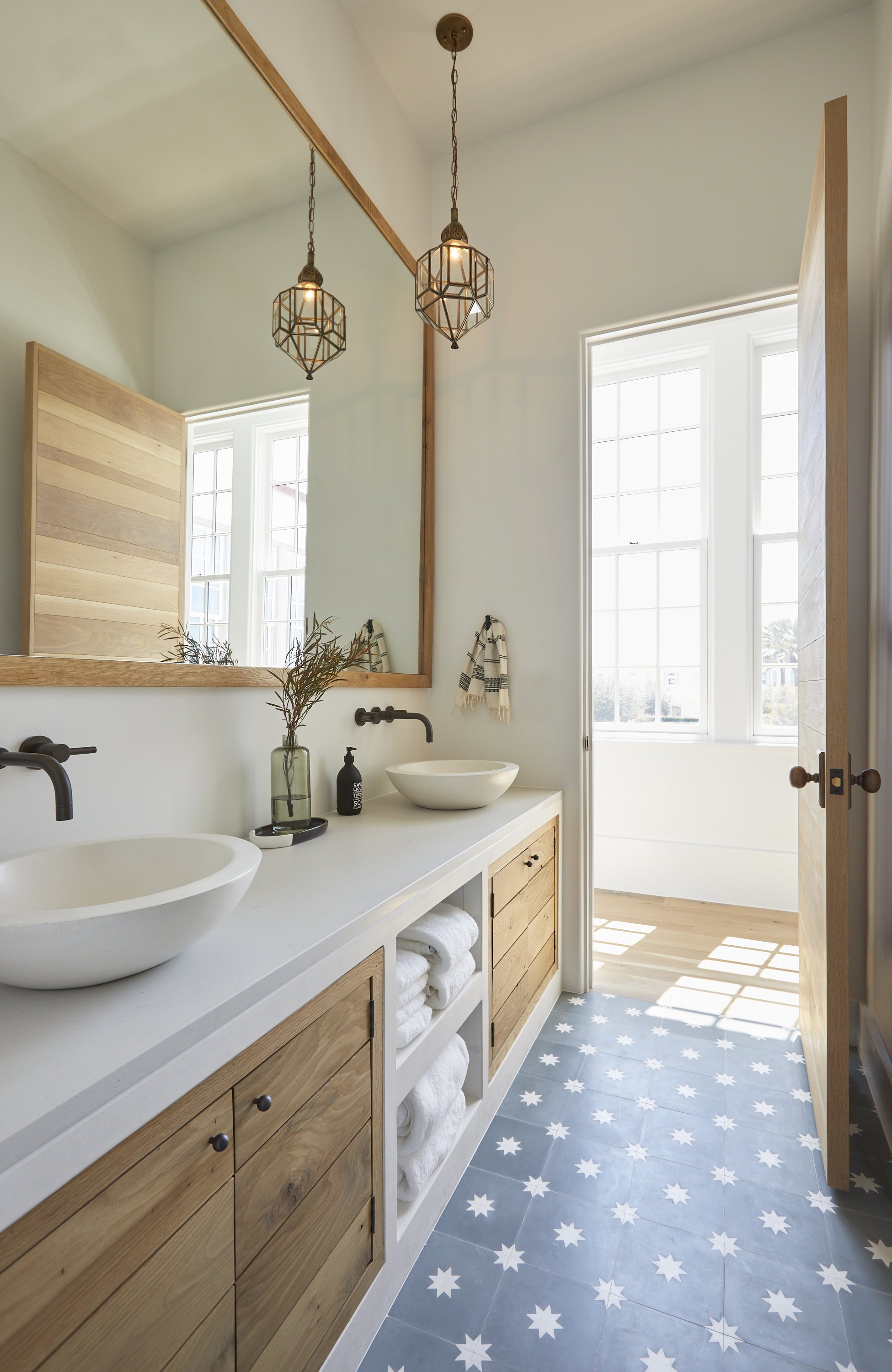 Modern bathroom with double sink vanity, wooden cabinets, large mirror, pendant lights, and large windows with sunlight streaming in, decorative black soap dispenser, towel hooks, and patterned floor tiles.