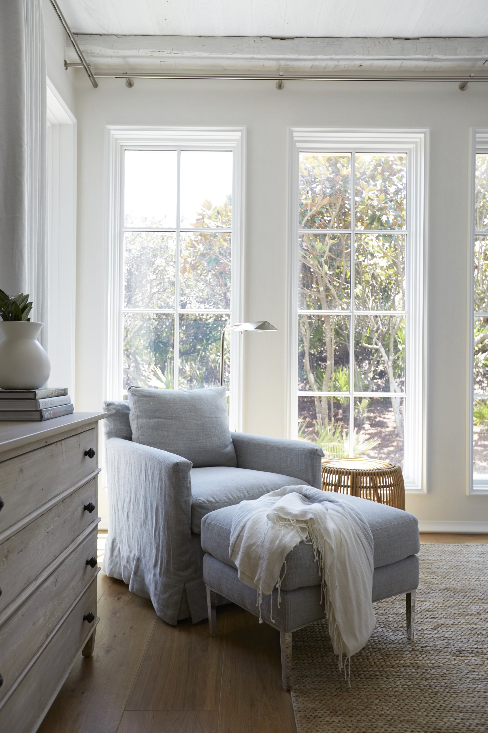 Sunlit living room with large windows, a gray armchair with a matching ottoman, a white throw blanket, a wooden side table, and a dresser with plants and books.
