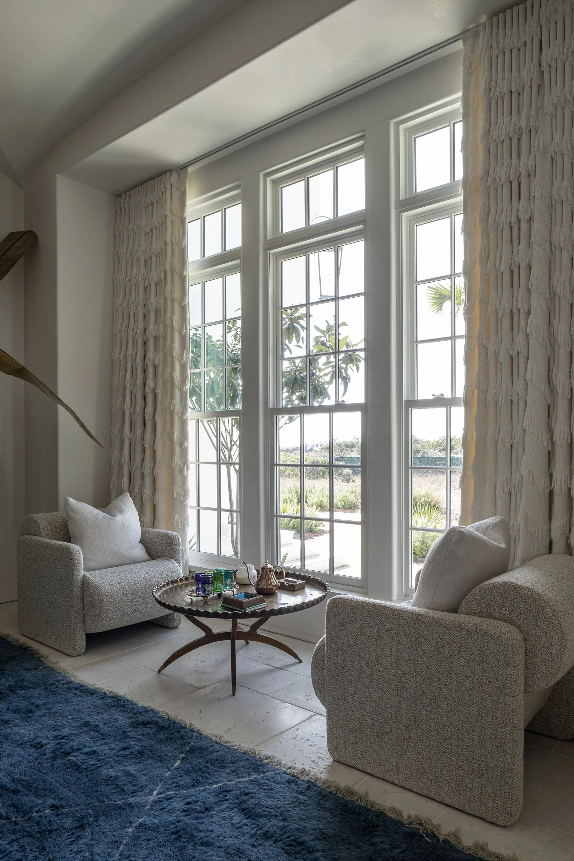 A living room with large windows, white curtains, two beige armchairs with pillows, a wooden coffee table with a tea set and glasses, a blue rug, and outdoor plants visible through the window.