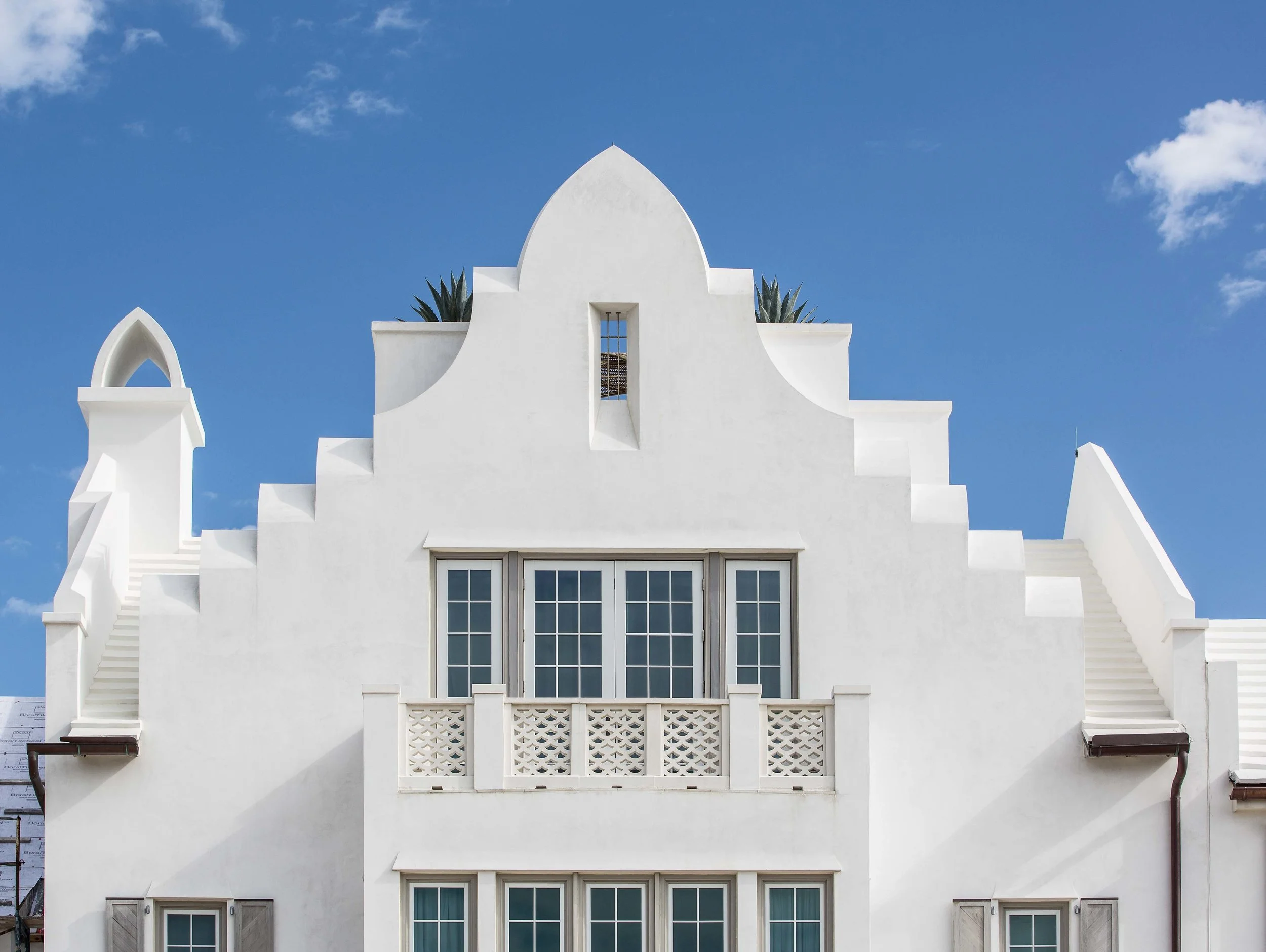 White building with architectural elements resembling a church or historic structure, featuring a large central window, decorative balcony, staircases on the sides, and a blue sky with scattered clouds in the background.