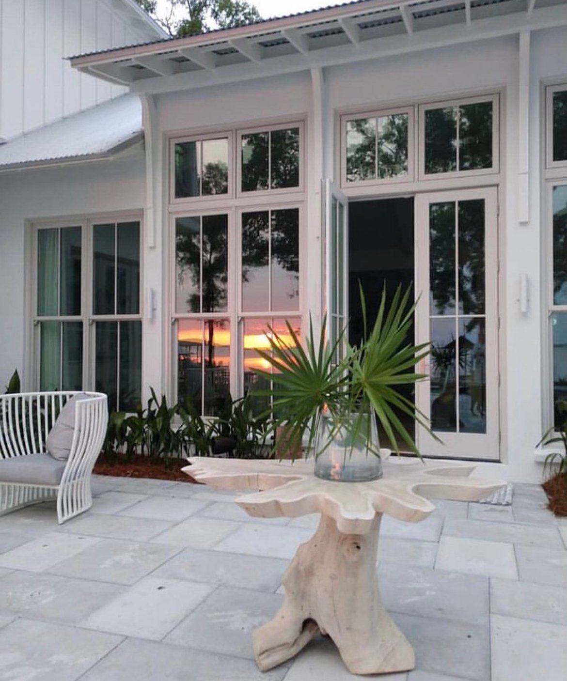 Patio with a wooden table, tropical plant in a glass vase, and outdoor furniture, with a sunset reflecting in large window panes of a modern house.