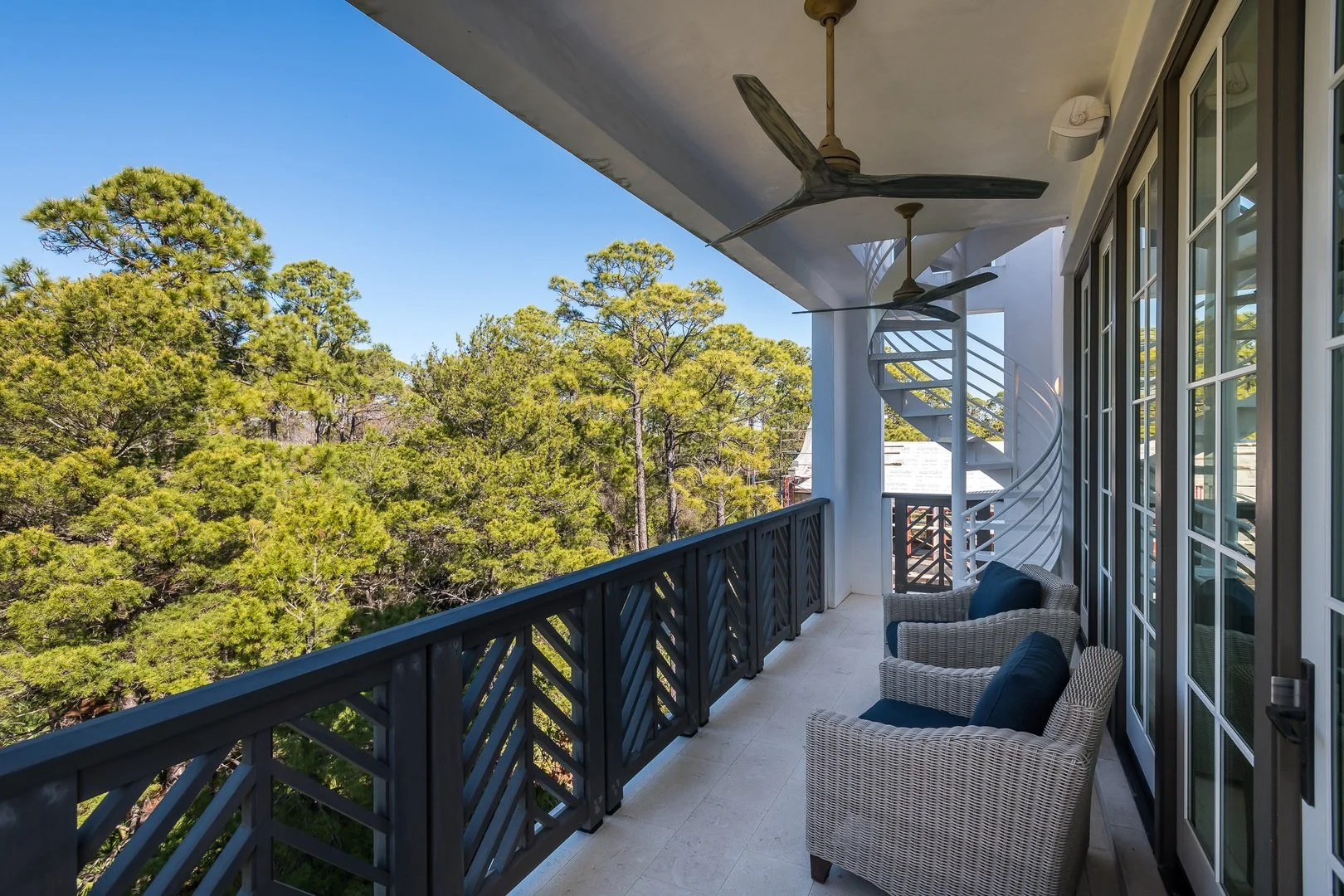 Balcony with two wicker chairs and navy cushions, black railing, and ceiling fans, overlooking green trees and a blue sky.