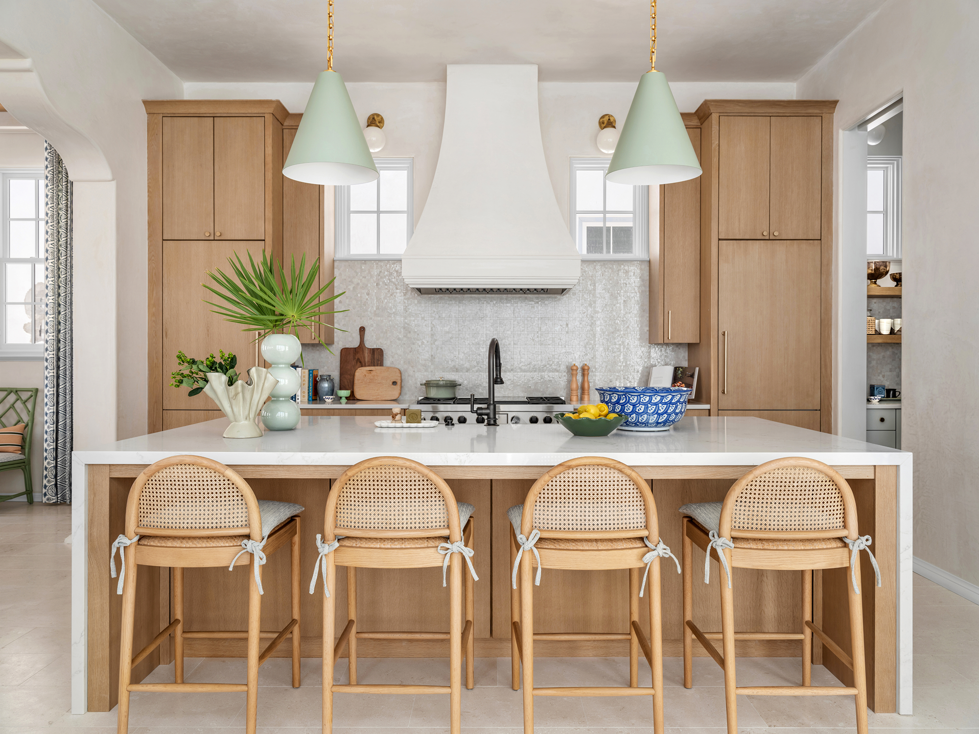 Modern kitchen with wooden cabinets, a white island, four wooden chairs tied with blue bows, potted plants, a blue patterned bowl with lemons, and large pendant lights.