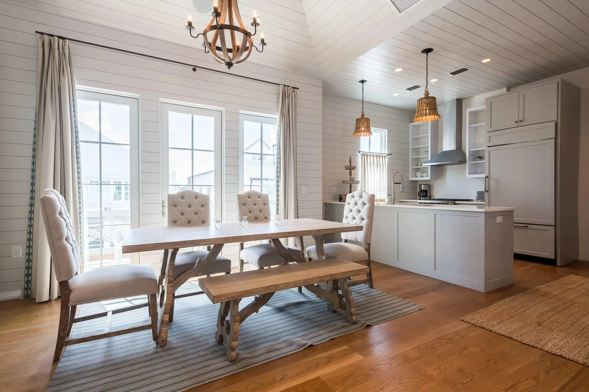 Bright dining area with wooden table, upholstered chairs, and sliding glass doors leading outside. Connected to a kitchen with white cabinetry, open shelving, and pendant lighting.